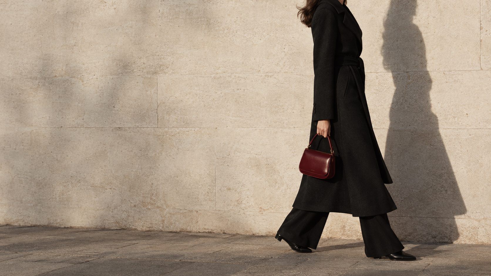 Mujer con abrigo negro y bolso color cereza caminando junto a un muro de piedra